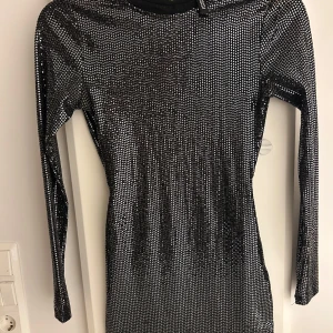 Glittrig svart klänning med öppen rygg - Snygg svart klänning med glittrigt mönster och en fantastisk öppen rygg. Perfekt för festliga tillfällen! Klänningen har långa ärmar och en tight passform som framhäver figuren. Ett måste för nyårsfesten eller en utekväll.