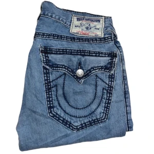 Blå jeans från True Religion - Säljer ett par snygga blå jeans från True Religion. De har en cool design med kontrasterande sömmar och klassiska fickor med knappar. Perfekta för en avslappnad stil. Passar både till vardags och fest! Priset är ej hugget i sten! 