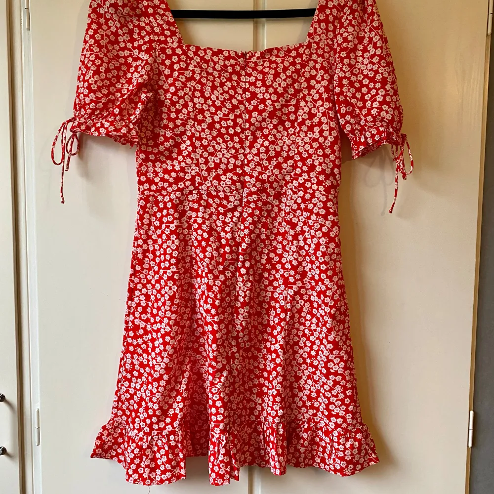 Superfin röd klänning med vita blommor. Den har korta ärmar med knytband och en söt volangkant längst ner. Klänningen är i ett lätt och luftigt material, vilket gör den idealisk för varma dagar. Passar både till vardags och festliga tillfällen.. Mekot.