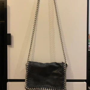 Svart axelremsväska från Tiamo - Snygg svart axelväska i skinn (lite sliten på framsidan) med coola kedjedetaljer runt kanterna. Väskan har en dragkedja på baksidan och stängs med magnetknappar. Perfekt för en kväll ute eller en dag på stan!