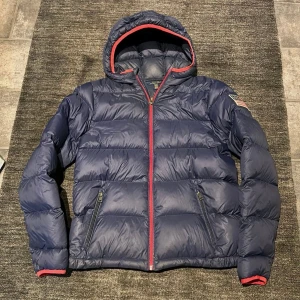 Blå dunjacka från Polo Ralph Lauren - En blå dunjacka med huva och röda detaljer. Jackan har en stor logotyp på baksidan och en mindre på bröstet. På vänster ärm sitter en liten amerikansk flagga. Fint begagnat skick, inga skador vad jag kan se. Storlek XL (18-20) motsvarar ca 170-176, S.  BVSA