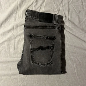 Nudie jeans - Säljer ett par grå jeans i bra skick med en cool svart detalj på bakfickan. De har en klassisk femficksdesign och en bekväm passform. Perfekta för både vardag och fest! 🖤