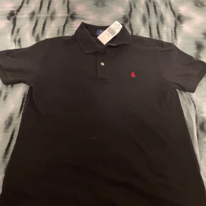 Svart pikétröja från Ralph Lauren - Säljer en snygg svart pikétröja från Ralph Lauren i slim fit. Tröjan är i nytt skick med etikett kvar och har en klassisk krage med knappar. Perfekt för både vardag och fest!                    Från ZALANDO FINNS KVITTO