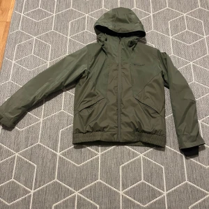 Grön vindjacka från Columbia - Säljer en grön vindjacka från Columbia i mycket bra skick. Jackan har en praktisk huva och dragkedja framtill. Perfekt för höst och vår, med en stilren design och normal passform. Den är långärmad och gjord i ett slitstarkt material.