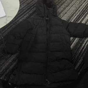 Svart dunjacka från McKinley - Säljer en svart dunjacka från McKinley i storlek 140. Jackan är perfekt för kalla vinterdagar med sin varma och vadderade design. Den har en praktisk huva och dragkedja framtill. Perfekt för att hålla sig varm och bekväm under vinterns äventyr! 🖤lång jacka 