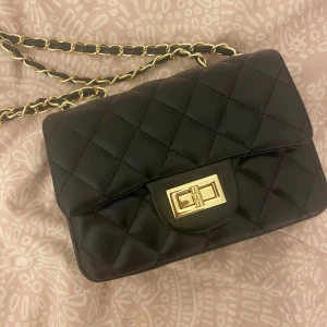 Svart quiltad axelväska med guldkedja - Snygg svart axelväska i quiltat mönster med guldfärgad kedja. Perfekt för både vardag och fest! Väskan är kompakt men rymmer det viktigaste. Kedjan ger en lyxig touch och quiltningen gör den extra trendig. Från glitter.