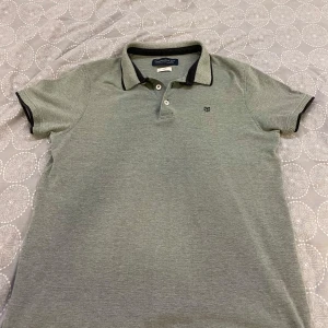 Grå pikétröja från Jack & Jones - Säljer en stilren grå pikétröja från Jack & Jones i storlek L. Tröjan har en klassisk krage med knappar och en liten logga på bröstet. Perfekt för både vardag och lite mer uppklädda tillfällen. Skön och enkel att matcha med det mesta! 🖤