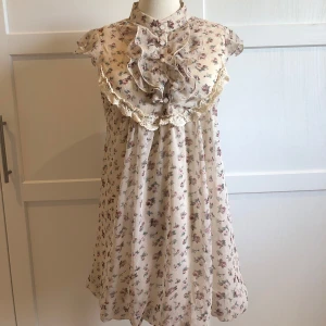 Blommig klänning från Lily J London - Söt blommig klänning från Lily J London i ljusa toner. Den har volangdetaljer och knappar framtill, med korta ärmar och en luftig passform. Perfekt för vår och sommar med sitt romantiska mönster och lätta material. Storleken står som M/L men den passar som S. Använd få gånger.