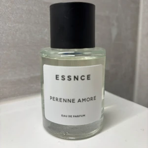 Essnce Perenne Amore Eau de Parfum - Säljer en elegant och stilren parfymflaska från Essnce med doften Perenne Amore. Flaskan är genomskinlig med en enkel vit etikett och svart kork. Perfekt för den som älskar en tidlös och sofistikerad doft. Passar både till vardags och fest!
