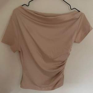 Säljer en snygg beige offshoulder topp med korta ärmar. Toppen har en cool rynkad detalj på sidan som ger den en unik look. 