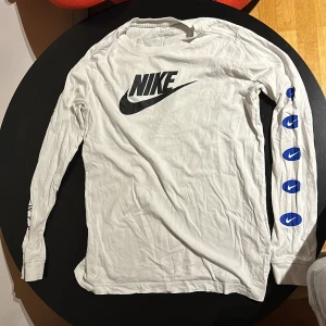Vit långärmad tröja från Nike - Säljer en vit långärmad tröja från Nike i bra skick. Den har det klassiska Nike-loggan i svart på bröstet och ryggen, samt flera blå Nike-loggor längs ena ärmen. Perfekt för en sportig och avslappnad stil. Passar bra till både vardag och träning.