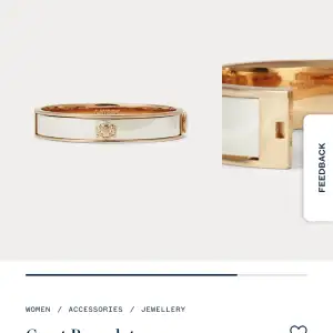 Snyggt och stilrent armband i guld med vit emaljdetalj. Armbandet har en elegant design med ett litet emblem i mitten. Perfekt för att ge en touch av lyx till vilken outfit som helst.