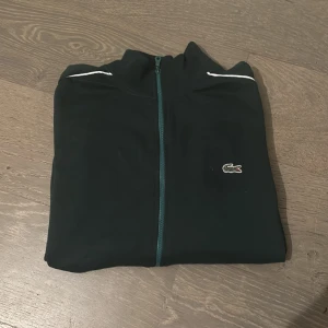 Svart tröja med dragkedja från Lacoste - Säljer en snygg svart tröja med dragkedja från Lacoste. Den har vita detaljer längs ärmarna och det klassiska krokodilmärket på bröstet. Perfekt för en casual look eller träning. Tröjan är i mycket bra skick och redo för nya äventyr!