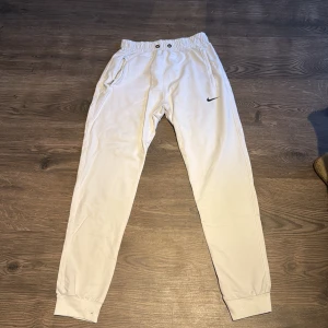 Vita mjukisbyxor från Nike - Säljer ett par vita mjukisbyxor från Nike i bra skick. De har en elastisk midja med snörning och muddar vid bensluten. Perfekta för en avslappnad stil eller träning. Byxorna har också fickor och den klassiska Nike-loggan på ena benet.