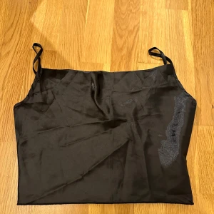 Svart axelbandslös tubtopp - Snygg svart axelbandslös tubtopp i ett slätt och glansigt material. Perfekt för en utekväll eller fest. Den har smala axelband som kan justeras.