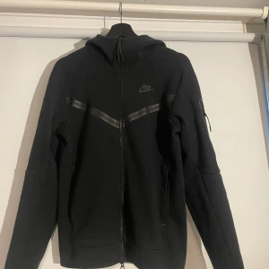 Svart tröja från Nike - Säljer en svart Nike-tröja i mycket bra skick. Den har en dragkedja framtill och en cool ficka med dragkedja på ärmen. Perfekt för höst och vår, med en stilren design och bekväm passform. Passar både till träning och vardagsbruk.