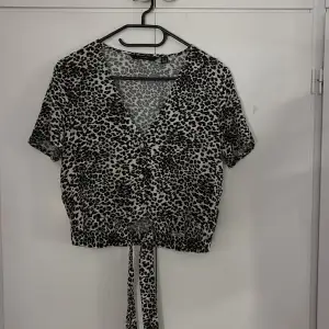 Säljer en snygg leopardmönstrad topp från Vero Moda i storlek M. Toppen har korta ärmar och en v-ringad design med knappar framtill. Den är croppad och har ett knytband i midjan för en extra detalj. Perfekt för en trendig look!