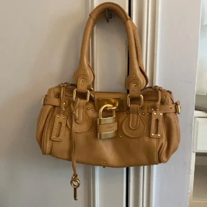 Beige handväska från Chloé -  (PRIS KAN DISKUTERAS VID SNABB AFFÄR)Snygg beige handväska från Chloé med gulddetaljer och ett stort hänglås framtill. Väskan har dubbla handtag och dekorativa nitar. Fint skick på väskan, finns en liten defekt på låset och en liten fläck på undersidan av väskan. Men inget som syns!🥰 