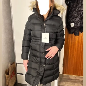 Svart Moncler dunjacka  - Snygg svart dunjacka från Moncler med pälsdetaljer på huvan. Jackan har en lång passform och stängs med dragkedja och knappar. Perfekt för kalla dagar. (Pälsen är utbytt mot äkta päls)