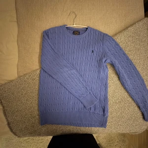 Blå stickad tröja från Ralph Lauren - Säljer en snygg blå stickad tröja från Ralph Lauren. Tröjan har ett klassiskt kabelstickat mönster och långa ärmar. Perfekt för kyligare dagar och ger en stilren look.