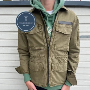 Olivgrön jacka med dragkedja - Snygg olivgrön jacka med dragkedja och flera praktiska fickor. Jackan har en cool militärinspirerad look med detaljer som axelklaffar och ärmficka. Perfekt för en avslappnad stil.