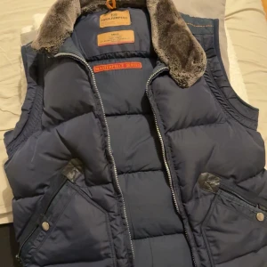 Blå dunväst från Parajumpers - Säljer en snygg blå dunväst från Parajumpers med fuskpäls på kragen och dragkedja framtill. Västen har broderad logga på bröstet och praktiska fickor med knappar. Perfekt för kyliga dagar! Och den är i bra skick och de svårt att hitta en mörkblå gammal modell. De går och diskutera om priset 
