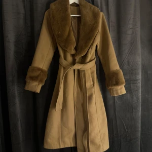 Beige kappa med pälsdetaljer - Elegant beige kappa med mjuka pälsdetaljer på krage och ärmslut. Kappan har ett knytbälte i midjan för en snygg siluett. Perfekt för kyliga dagar när du vill hålla stilen.