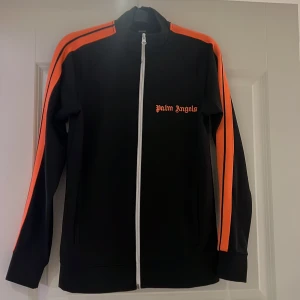 Svart ziptröja från Palm Angels - Snygg svart ziptröja från Palm Angels med orangea ränder längs ärmarna och dragkedja framtill. Jackan har en hög krage och är långärmad, perfekt för en sportig look. I storlek S