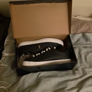 Svarta sneakers från Polo Ralph Lauren - Snygga svarta sneakers från Polo Ralph Lauren med vita snören och en vit sula. Skorna har en ikonisk logga på sidan och är perfekta för en stilren look.