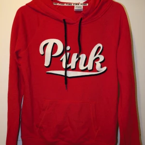 Röd hoodie från Victoria's Secret Pink - Säljer en snygg röd hoodie från Victoria's Secret Pink med stor vit text på framsidan. Den har en känguruficka och svarta dragsnören vid huvan. Perfekt för en avslappnad stil!