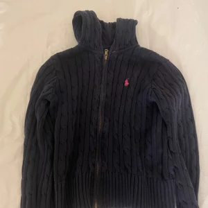 Svart stickad hoodie från Ralph Lauren - Säljer en snygg svart stickad hoodie från Ralph Lauren med dragkedja och det klassiska rosa logotypen på bröstet. Perfekt för kyliga dagar med sin mysiga huva och ribbade detaljer vid ärmslut och nederkant.