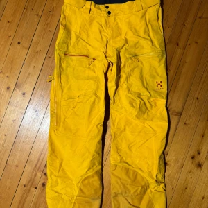 Gula GoreTex-byxor från Haglöfs - Säljer ett par gula skalbyxor i trelagers GoreTex från Haglöfs, modell ROC/VOJD. Byxorna har praktiska dragkedjor och är tillverkade i ett slitstarkt material, perfekt för utomhusaktiviteter som skid- och Snowboardåkning. Plaggens ljust gula färg har fläckats något av svarta vantar i skinn men det påverkar givetvis inte funktionen. 