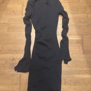 Svart ribbad klänning - Snygg svart ribbad klänning med lång ärm och asymmetrisk hals. Perfekt för en stilren look. Klänningen har en tight passform som framhäver figuren.