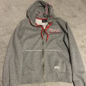 Grå zip hoodie från Peak Performance - Säljer en grå hoodie från Peak Performance med röd text och dragkedja. Den har en justerbar huva med röda snören, storlek L men tycker den är lite liten för att vara L. Säljer för den är för stor för mig☺️