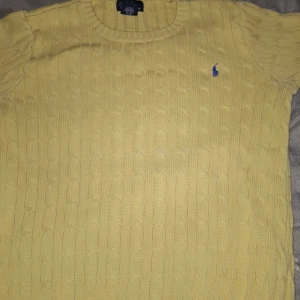 Gul stickad tröja från Ralph Lauren - Säljer en gul stickad tröja från Ralph Lauren med kabelstickat mönster och en blå logga på bröstet. Tröjan har korta ärmar och rund halsringning. Perfekt för en avslappnad stil. Strl xs - s