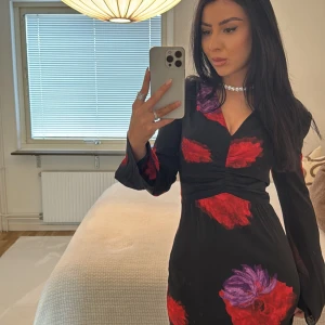 Svart långklänning med blommönster från ADOORE - Elegant svart långklänning från ADOORE med röda och lila blommor. Klänningen har en djup v-ringning och långa ärmar. Den har en slits framtill som ger en stilfull touch.36 size