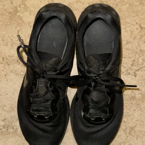 Svarta Nike sneakers - Säljer ett par svarta Nike sneakers med snörning. Skorna har en stilren design med en svart sula och Nike-logga på sidan. Perfekta för vardagsbruk och träning.