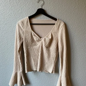 Beige ribbad topp med volangärmar - Snygg beige ribbad topp med långa volangärmar och en söt knytdetalj framtill. Perfekt för en stilren look.