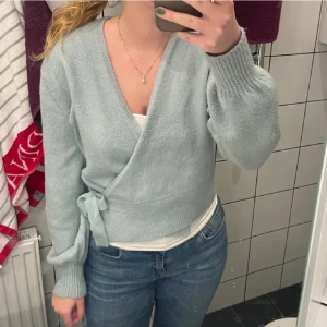 Ljusblå omlottkofta - Mysig ljusblå omlottkofta med knytband i midjan. Tröjan har långa ärmar och ribbade muddar, perfekt för en avslappnad stil. Passar bra till jeans eller kjol.