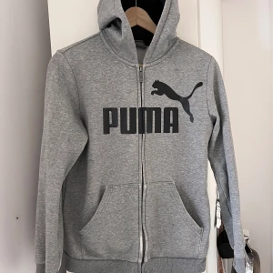 Grå hoodie från Puma storlek 164 - Säljer en grå hoodie från Puma med dragkedja. Tröjan har långa ärmar och en praktisk huva. Den  är lite nött på sina ställen. Se bilder. Inga hål, fläckar.. 
