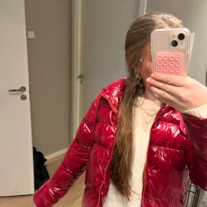 Röd pufferjacka från Guess - Snygg röd pufferjacka från Guess med glansig finish och dragkedja framtill. Jackan har en huva och är perfekt för kyligare dagar. Den är i storlek XS och är väldigt trendig, samt slutsåld på hemsidan.
