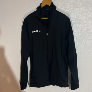 Svart långärmad jacka från Craft - Säljer en svart långärmad jacka från Craft i storlek XL. Jackan har en dragkedja framtill och är perfekt för träning. Den är tillverkad i ett syntetmaterial som ger bra komfort och rörelsefrihet.
