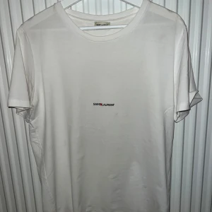 Saint Laurent tshirt - Säljer en stilren vit t-shirt från Saint Laurent med diskret logga på bröstet. Perfekt för en minimalistisk look. T-shirten har en klassisk rund hals och korta ärmar. Kom privat vid fler frågor, tar bud och trades! 