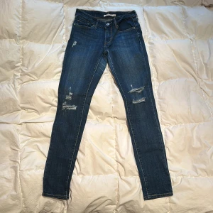 Blå skinny jeans från Levi's - Säljer ett par snygga blå skinny jeans från Levi's med slitna detaljer. De har en klassisk femficksdesign och knappgylf.