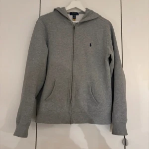 Ralph lauren zip  - Säljer en stilren grå hoodie från Ralph Lauren med dragkedja och broderad logga på bröstet. Tröjan har långa ärmar och ribbade muddar, perfekt för en avslappnad look. Passar bra till både jeans och joggers. Bara använd några gånger. Bra nu när det är kallt ute. Passar perfekt till vardagliga användningar och även till finare tillfällen. Har bara fått ett litet snitt som man ser på bilden. Storlek 16 år i barn passar som S i herr.