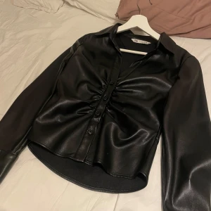 Svart skjorta från Zara - Snygg svart skjorta från Zara i skinnimitation med knappar framtill och rynkdetaljer. Perfekt för en trendig look.