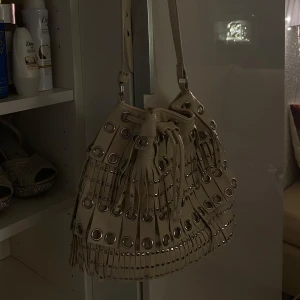 Stockhol stil Beige väska med nitar  - Snygg beige axelväska med coola nitar och fransdetaljer. Väskan har en justerbar axelrem och dragsko för stängning. Perfekt för att ge din outfit en edgy touch.