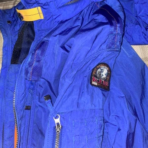Blå jacka från Parajumpers - Säljer en blå jacka från Parajumpers med dragkedja och flera fickor. Jackan har en gul detalj vid kragen och en patch på ärmen. Perfekt för höst och vår.