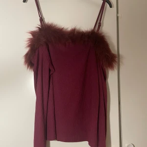 Vinröd topp med fuskpälsdetalj - Snygg vinröd topp med fuskpälsdetalj längs halsringningen. Toppen har smala axelband och långa ärmar, vilket ger en elegant look. Perfekt för en kväll ute eller en speciell tillställning.