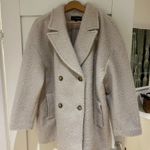 Beige teddyjacka från KappAhl - Mysig beige teddyjacka från KappAhl med dubbelknäppning och stora slag. Perfekt för kyliga dagar med sin fluffiga textur och stilrena design. Jackan har två fickor framtill och en bekväm passform.